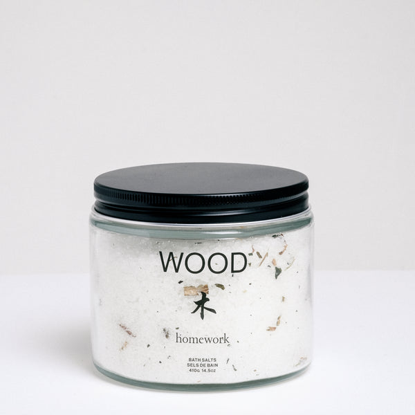 A short, cylindrical glass jar of bath salts with a flat, black, metal screwtop lid. The jar has WOOD written on it, and the Japanese kanji for wood, with "homework" written below. Available at NiMi Projects UK.