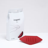 A red Japanese Irogami stainless steel grater in front of a packaged grater. The white paper packaging is illustrated with a line drawing of how to use the grater. Made in Japan and available at NiMi Projects. 