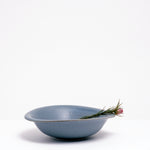 A side view of a Mino-yaki Japanese porcelain breakfast bowl, featuring an asymmetric curved rim and soft blue, flecked glaze and holding a sprig of flowers. On show at NiMi Projects UK.