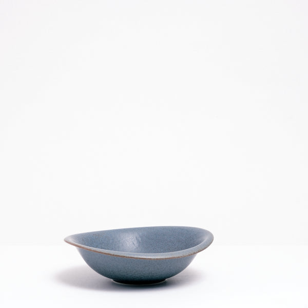 A side view of a Mino-yaki Japanese porcelain dessert bowl, featuring an asymmetric curved rim and soft blue, flecked glaze. On show at NiMi Projects UK.