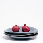 Three stacked Japanese Mino-yaki porcelain Flare plates, each with asymmetric curved rims and glazed in a soft blue glaze featuring tiny flecks. Two pomegranates are placed on top of the stack of plates, on show at NiMi Projects UK.
