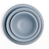 Three stacked Japanese Mino-yaki porcelain Flare plates and bowls, each with asymmetric curved rims and glazed in a soft blue glaze featuring tiny flecks. On show at NiMi Projects UK.
