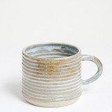 A ribbed stoneware mugs by Maki Baxter with warm mint and beige glaze and exposed clay base. Available at NiMi Projects UK.