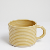 A yellow glazed ribbed stoneware mug by Maki Baxter with an exposed clay base.  Available at NiMi Projects UK.