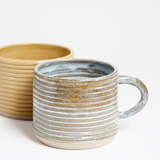 Two ribbed stoneware mugs by Maki Baxter with warm glaze and exposed clay base. One in a yellow glaze, the other in a mint and beige glaze. Available at NiMi Projects UK,