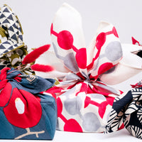 A group of gifts, wrapped in various furoshiki wrapping cloths by NiMi Projects UK. From left to right — a green gourd furoshiki, a blue tsubaki (camellia) furoshki, a grey and white dotted furoshiki and a blue and white furoshiki with pine motifs.