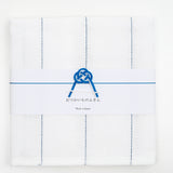 Ariel view of a white, folded Japanese fukin kitchen cloth, detailed with blue stitched vertical lines and wrapped in a white slip with a blue and white mizuhiki paper cord knot in a traditional design mount fuji. Available at NiMi Objects.