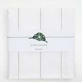 Top view of a white, folded Japanese fukin kitchen cloth, detailed with green vertical stitched lines and wrapped in a white slip with a green and white mizuhiki paper cord knot in a traditional pine design. Available at NiMi Objects.