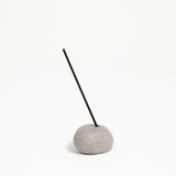 A grey Maki Baxter ceramic Incense Pebble Incense holder with stick of dark coloured Japanese incense. Behind are two white versions. Available at NiMi Projects UK.
