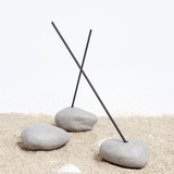 Three grey Maki Baxter ceramic Japanese Incense Pebble Incense holders standing on a bed of sand. Two of the pebbles are holding dark sticks of incense. On show at NiMi Projects UK.
