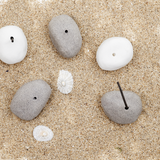 A group of ceramicist Maki Baxter's Pebble Japanese Incense Holders in white and grey, on a background of sand and sea shells. Available at NiMi Projects UK.