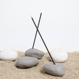 Three grey Maki Baxter ceramic Japanese Incense Pebble Incense holders, two of which are holding dark sticks of incense. Behind are two white versions. On show at NiMi Projects UK.
