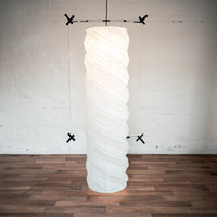 A 130-centimeter tall Saiko Design Neji Neji lampshade on show at NiMi Projects UK. The lampshade is a tall gently twisted cylindrical form with curved ridges. Its framework winds around the shape and is made using traditional chochin Japanese lantern techniques.