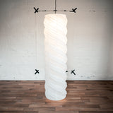 A 130-centimeter tall Saiko Design Neji Neji lampshade on show at NiMi Projects UK. The lampshade is a tall gently twisted cylindrical form with curved ridges. Its framework winds around the shape and is made using traditional chochin Japanese lantern techniques.