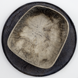 Top view of a rectangular Chinaberry wood bowl, showing the texture of its hand-chisel carving, and finished in urushi silvery grey lacquer with a shimmery dusting of fine tin powder. The bowl is placed on top of a round wooden plate textured with layers of washi paper and deep bronze lacquer. Made by Solmu Puut in Japan from wood collected in Yakushima and on display at NiMi Projects UK.