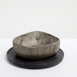 Side iew of a rectangular Chinaberry wood bowl, showing the texture of its hand-chisel carving, and finished in urushi silvery grey lacquer with a shimmery dusting of fine tin powder. The bowl is placed on top of a round wooden plate textured with layers of washi paper and deep bronze lacquer. Made by Solmu Puut in Japan from wood collected in Yakushima and on display at NiMi Projects UK.