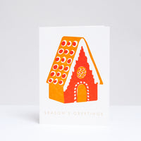 Takako Copeland's Gingerbread House greeting card, featuring a letterpress-printed cinnamon orange and red gingerbread house with and the words “Season’s Greetings” in gold foil. Available at NiMi Projects UK.