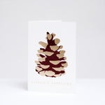Takako Copeland's Christmas Gold Pine Cone greeting card, featuring a letterpress-printed aubergine-coloured pine cone with gold highlights and the words “Season’s Greetings” in gold foil.