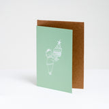 A pistachio green greeting card at NiMi Projects UK, featuring a white line drawing design by Takako Copeland of a cat wearing a jumper and party crown and holding a platter with a large trifle topped with a star. Inside the card is a natural brown envelope.