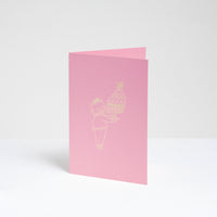 A standing pink greeting card, designed by Takako Copeland, featuring a gold foil line drawing illustration of a cat in a party crown and sweater holding a large trifle on a platter. Available at NiMi Projects UK. 