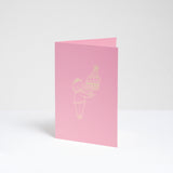 A standing pink greeting card, designed by Takako Copeland, featuring a gold foil line drawing illustration of a cat in a party crown and sweater holding a large trifle on a platter. Available at NiMi Projects UK. 