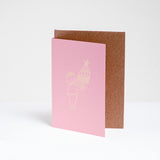 A standing pinke greeting card, designed by Takako Copeland, featuring a gold foil line drawing illustration of a cat in a party crown and sweater holding a large trifle on a platter. Available at NiMi Projects UK. 