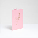 A standing pink greeting card, designed by Takako Copeland, featuring a gold foil line drawing illustration of a cat in a party crown and sweater holding a large trifle on a platter. Available at NiMi Projects UK. 