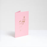 A standing pink greeting card, designed by Takako Copeland, featuring a gold foil line drawing illustration of a cat in a party crown and sweater holding a large trifle on a platter. Available at NiMi Projects UK. 