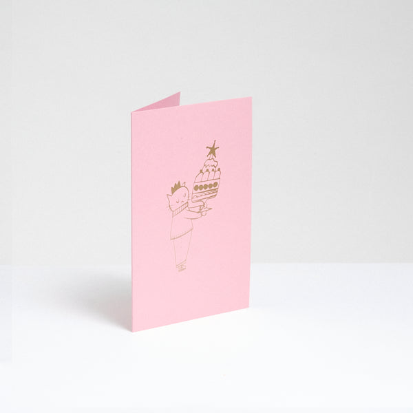 A standing pink greeting card, designed by Takako Copeland, featuring a gold foil line drawing illustration of a cat in a party crown and sweater holding a large trifle on a platter. Available at NiMi Projects UK. 