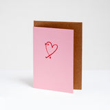 A standing pink card with a red line drawing illustration of a bird shaped as a heart by Takako Copeland. Inside the card is a natural brown envelope. On show at NiMi Projects UK.