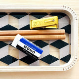 A styled flat-lay of stationery items arranged on a geometric tray. The set includes a Japanese gold metal SUN clip, two pencils, and a white Japanese Classic Mono Eraser with its blue and black sleeve. NiMi Projects UK.