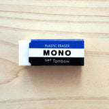 Tombow MONO Classic block eraser in its  iconic blue, white and black striped paper sleeve, shown on a wood background. Made in Japan and available at NiMi Projects UK.
