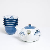 A side view of a vintage white porcelain yokode (side-handled) Japanese teapot. Next to the teapot are five stacked teacups. The teapot features a blue lid and a hand-painted blue design of a mountainscape, while the stacked cups have blue exteriors and mountains painted inside. On show at NiMi Projects UK.
