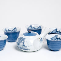 A vintage white porcelain Japanese tea set, including a tea yokode (side-handled) teapot and five teacups. The teapot features a blue lid and a hand-painted blue design of a mountainscape, while the cups are blue with mountains painted inside. Available at NiMi Projects UK.
