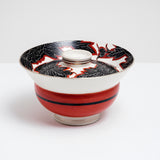 Side view of a white vintage, Japanese porcelain, decorative lidded donburi bowl, showing a bold pattern of peonies in black with white detailing and brick red borders on the lid and a band of brick red on the side. On show at NiMi Projects UK.
