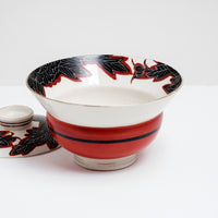 Side view of a white vintage, Japanese porcelain, decorative lidded donburi bowl, showing a bold pattern of peonies in black with white detailing and brick red borders on the rim and a band of brick red on the side. On show at NiMi Projects UK.