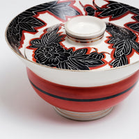 Close up of a vintage, Japanese porcelain, decorative lidded donburi bowl, showing a bold pattern of peonies in black with white detailing and brick red borders on the lid and a band of brick red on the side. On show at NiMi Projects UK.