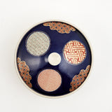 Top view of a Japanese, vintage, round porcelain lidded bowl showing a deep blue glaze, three circular traditional patterned motifs in brick red, gold and pale blue and three cloud-shaped patterned motifs in red and gold the edges.