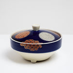 Side view of a Japanese, vintage, round porcelain lidded bowl showing a deep blue glaze, three circular traditional patterned motifs in brick red, gold and pale blue and three cloud-shaped patterned motifs in red and gold the edges.