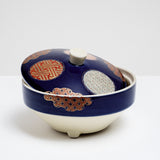 A Japanese, vintage, round porcelain lidded bowl showing a deep blue glaze, three circular traditional patterned motifs in brick red, gold and pale blue and three cloud-shaped patterned motifs in red and gold the edges.