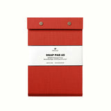 A Signal Red fabric-covered A4 Postalco Snap Pad, showing the two roundmetal snap clasps at its top and a white label wrapped around its center featuring the Postalco bird logo.