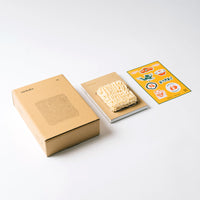 The Sankaku Vol. 02: Ramen book limited edition set. From left to right: The limite-edition cardboard box with an illustration of ramen noodles by Stanley Sun; the hardback book itself, featuring vacuu-packed real instant noodles as part of its cover; and a sheet of stickers of cute illustrations relating to ramen.