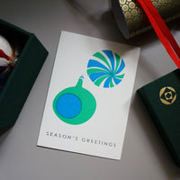 A letterpress printed Christmas card, designed by Takako Copeland, featuring two baubles in green and blue on a white background, and gold lettering spelling out "Season's Greetings." The card is on display with an arrangment of festive decorations.