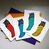 A group of four Takako Copeland illustrated and letterpress printed Christmas cards, featuring black cats in different coloured knitted stockings. From left to right: a black cat in a red stocking, blue stocking, green stocking and yellow stocking.
