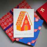 A letterpress printed Christmas card, designed by Takako Copeland, featuring a Gingerbread house in cinnamon orange and red, and the words "Season's Greetings” in gold foil. The card is on display with an arrangement of colourfully wrapped Christmas gifts.