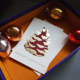 A letterpress printed Christmas card, designed by Takako Copeland, featuring an aubergine-coloured pine cone with gold-foil highlights and the words "Season's Greetings” in gold. The card is on display with an arrangement of festive baubles.