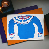 Takako Copeland’s Icelandic Sweater letterpress-printed card features an illustration of a bright blue and red patterned sweater that spreads across both sides of the card. It is displayed laid flat with brown and purple envelopes behind it and an arrangement of Christmas baubles.