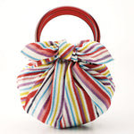 A multicolored striped furoshiki wrapping cloth tied into a pouch shaped bag with tortoise-shell colored ring handles, made in Japan and available at NiMi Projects UK.