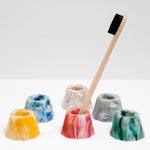 A collection of NiMi Projects’ six Buoy recycled marine plastic toothbrush stands, in yellow blue, white, grey, green and red. The red toothbrush stand sits in the middle and holds a bamboo toothbrush. 