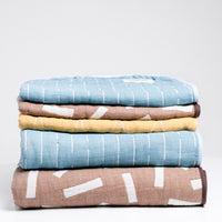 A stack of Blanked Japanese cotton gauze blankets on display at NiMi Projects UK. From top to bottom: a blue small blanket with white line patterning, a small brown version with white rectangles, a small yellow version with white circles, a large blue blanket and a large brown blanket.